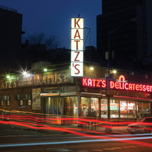 Katz's Delicatessen at night, the lit KATZ'S tower sign above the storefront with light trails across the street